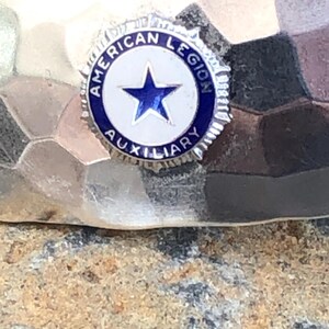 May include: A silver-toned pin with the words "AMERICAN LEGION AUXILIARY" encircling a white star on a blue background. The pin is set against a textured, hammered metal surface.