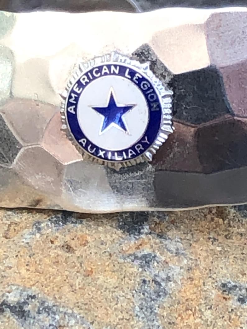 May include: A silver-toned pin with the words "AMERICAN LEGION AUXILIARY" encircling a white star on a blue background. The pin is set against a textured, hammered metal surface.