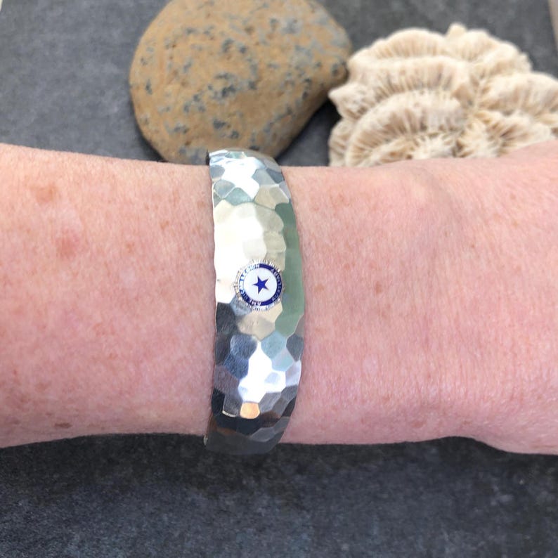 May include: A silver hammered metal bracelet with a blue and white emblem featuring a star. The bracelet is worn on a wrist, showcasing its textured surface and design. The bracelet is approximately 2.5 inches wide.