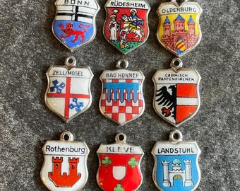Op de afbeelding: Een verzameling van negen schildvormige metalen bedels met kleurrijke emaille ontwerpen en de namen van Duitse steden, waaronder Bonn, Rüdesheim, Oldenburg, Zell/Mosel, Bad Honnef, Garmisch-Partenkirchen, Rothenburg, Kleve en Landstuhl.