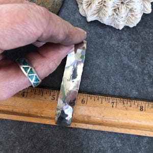 May include: A hammered silver cuff bracelet with a textured surface. The bracelet is held near a ring with turquoise inlay. A wooden ruler with inch markings is in the foreground.