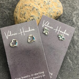 May include: Two pairs of sterling silver stud earrings on gray cards, one heart-shaped with blue accents, the other a flower design. The cards read "Valerie Hawkins" and "Fine jewelry in sterling silver and art jewelry in mixed metals."