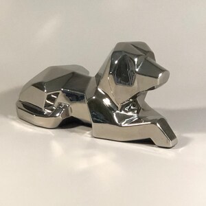 May include: A silver, geometric, stylized dog sculpture. The dog is lying down with its head turned to the side.