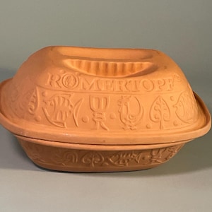 May include: A terracotta Romertopf clay baker with a lid, featuring embossed designs of fish, roosters, and leaves around the exterior. The word ROMERTOPF is clearly stamped on the top center of the lid, which includes a recessed handle.