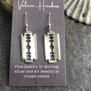 May include: A pair of silver-colored earrings shaped like razor blades. The earrings hang from silver hooks with small, decorative beads. The earrings are displayed on a gray card with the text "Valerie Hawkins" and "Fine jewelry in sterling silver and art jewelry in mixed metals."