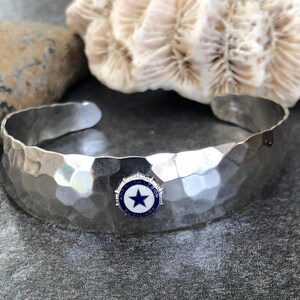 May include: Hammered silver cuff bracelet with a blue and white emblem featuring a star. The bracelet has a textured surface and is displayed on a dark gray surface with a rock and coral in the background.