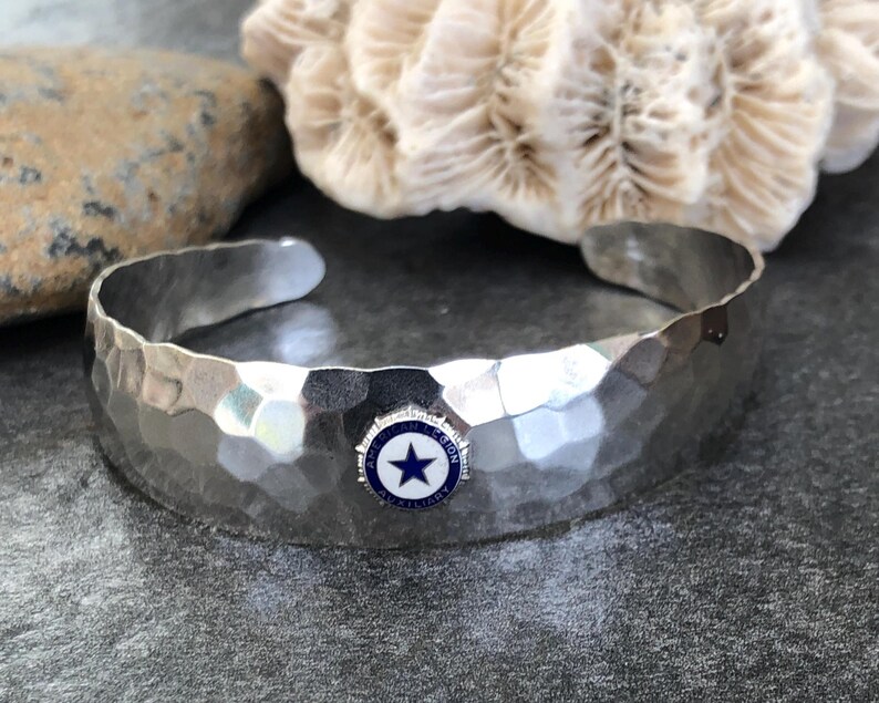 May include: Hammered silver cuff bracelet with a blue and white emblem featuring a star. The bracelet has a textured surface and is displayed on a dark gray surface with a rock and coral in the background.