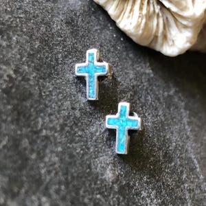 Peut inclure: Une paire de boucles d'oreilles clous en forme de croix en argent avec incrustation turquoise. Les boucles d'oreilles sont petites et détaillées, avec une couleur bleu vif. Les croix sont posées sur un fond sombre et texturé.