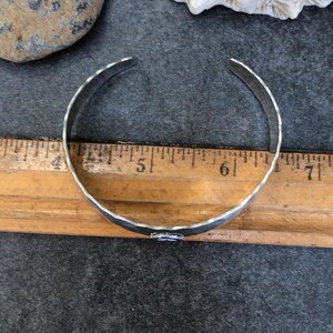 May include: A silver-colored cuff bracelet with a hammered texture. The bracelet is open-ended and measures approximately 6 inches in circumference. The bracelet is resting on a wooden ruler.