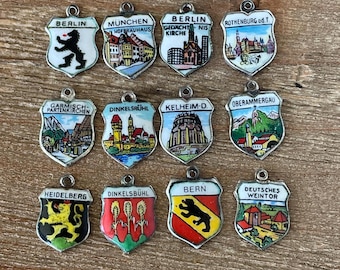 May include: A collection of twelve shield-shaped souvenir charms, each with a different city name and emblem. The charms feature colorful enamel designs, including buildings, landmarks, and coats of arms. The charms are made of metal and have a small loop at the top.