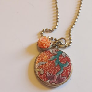 May include: A silver-toned ball chain necklace featuring a circular pendant with a red, teal, and white floral pattern. Above the circular pendant sits a small, light peach-colored carved flower charm attached to the same silver-toned metal bail.