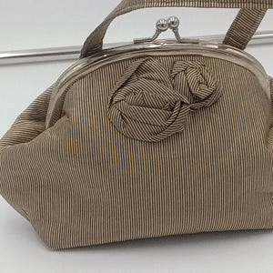 May include: A vintage-style handbag with a striped pattern in shades of brown and beige. The bag features a metal clasp closure and a small handle. A fabric flower adorns the front. The bag is approximately 20 cm wide.