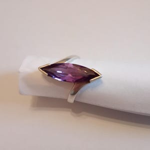 May include: A silver ring featuring a marquise-cut, deep purple gemstone. The gemstone is set in a gold bezel, and the ring has a simple, elegant design. The ring is displayed on a white surface, showcasing its color and shape.