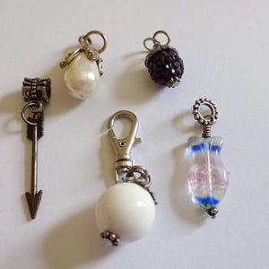 May include: A collection of decorative charms and pendants. Includes a pearl-shaped charm, a dark purple berry-shaped charm, a clear glass charm with blue and pink accents, a metal arrow charm, and a white ball charm with a clasp.