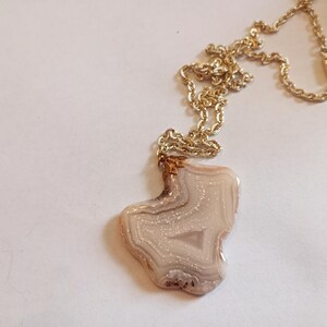 Peut inclure: Un collier doré avec un pendentif en pierre claire et ornée. La pierre a une forme unique avec des nuances de blanc, beige et marron. La chaîne est de conception torsadée et délicate.
