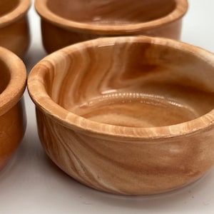 May include: Four small, round bowls are shown in a close-up. They are a warm, earthy brown with a marbled effect. The rims are a slightly lighter shade. The bowls are empty and seem to be made of ceramic or a similar material.