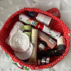 May include: A red polka-dot cosmetic bag filled with various beauty products. The bag contains bottles, tubes, and jars of skincare and makeup items. Two white crocheted face scrubbies are visible. The bag is on a white surface.