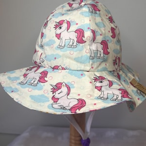 May include: A cream-coloured sun hat with a repeating pattern of pink-maned unicorns, blue clouds, and pink stars. The hat has a wide brim and a chin strap with a purple toggle. The hat is designed for children.