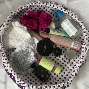 May include: A white and purple polka dot drawstring bag filled with various beauty products. Items include a pink scrunchie, small bottles, makeup compacts, and a white crocheted pouch. The bag is on a white, textured surface.