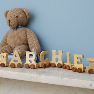 May include: A wooden toy train with the name ARCHIE spelled out in individual wooden letters. The train is brown and the letters are natural wood color. A brown teddy bear sits behind the train.