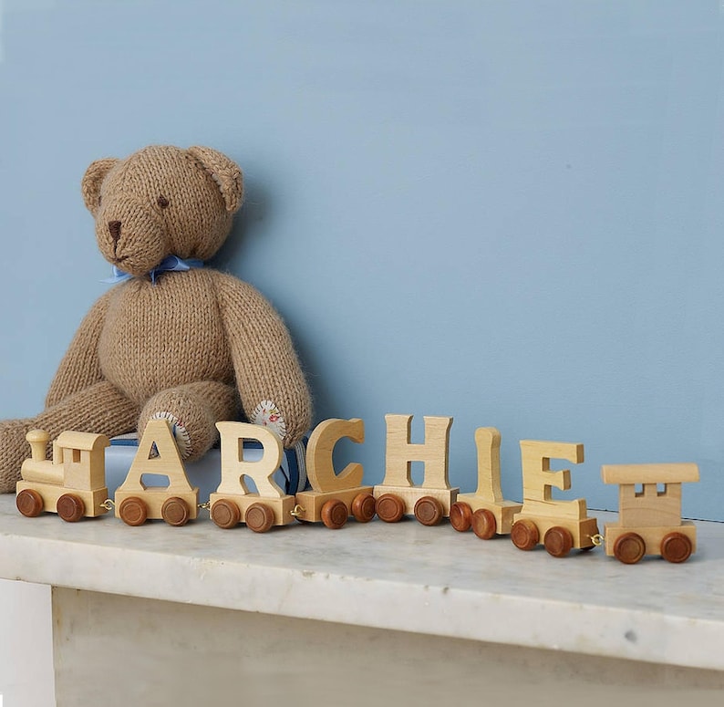 May include: A wooden toy train with the name ARCHIE spelled out in individual train cars. The train is on a white surface with a brown teddy bear sitting behind it.