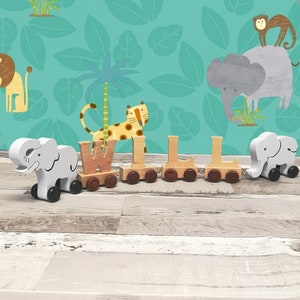 May include: A wooden toy train spelling "WILL". The train cars are letter-shaped and linked together. Elephant-shaped carriages are at each end. The background shows a jungle-themed wallpaper with illustrations of animals and foliage.