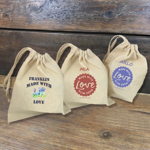 May include: Three small, beige drawstring bags with personalized designs. The first bag has a blue train with the text "Franklin Made With Love". The second bag has a red heart with the text "Made With Love" and the name "Mia". The third bag has a purple circle with the text "Made With Love" and the name "Arlo".