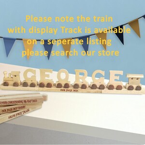 May include: Wooden train with the name "GEORGE" spelled out in individual train cars. The train is on a wooden track with a sign that says "OUR PAGE BOY". The train is decorated with brown and red accents. There are three wooden blocks below the train with the text "YOUR CHRISTENING 5th MAY 2023", "OUR PAGE BOY HARRY", and "ARLOS FIRST TRAIN".