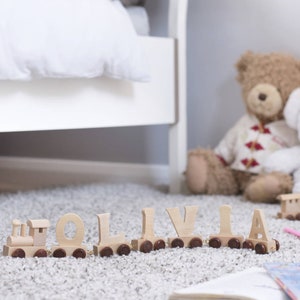 May include: Wooden train with the name Olivia spelled out in individual letters. The train is on a light brown carpet with a teddy bear and a book in the background.