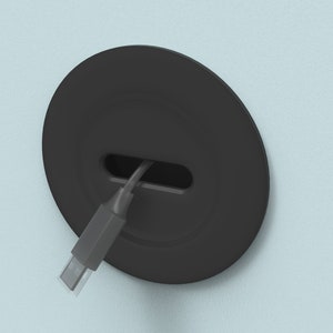 Puede incluir: Una placa de pared redonda negra con un cable que pasa por una ranura. El cable es un cable USB gris.