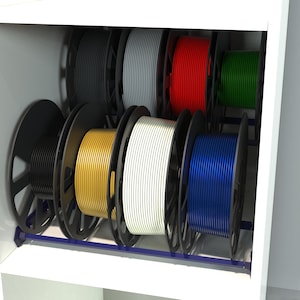 Può includere: Uno scaffale con un rack che contiene bobine di filamento per una stampante 3D. Le bobine sono di vari colori, tra cui nero, grigio, rosso, verde, oro, bianco e blu.