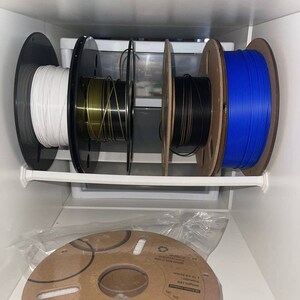May include: Four spools of filament for a 3D printer are organized on a shelf. The spools are white, gold, black, and blue. The spools are labeled with the text "Weight:1KG Diameter: 3.75 0.02 spool box made from recycled material".