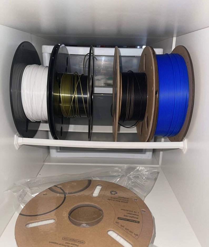 May include: Four spools of filament for a 3D printer are organized on a shelf. The spools are white, gold, black, and blue. The spools are labeled with the text "Weight:1KG Diameter: 3.75 0.02 spool box made from recycled material".
