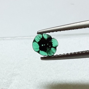 May include: A green emerald gemstone with black inclusions, held by tweezers. The emerald is cut into a flower shape.