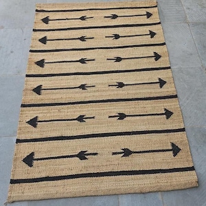 Op de afbeelding: Rechthoekig jute vloerkleed met een natuurlijke bruine achtergrond. Het kleed heeft horizontale zwarte strepen met pijlen die in afwisselende richtingen wijzen. Geschikt voor binnenshuis.