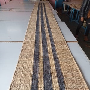 Puede incluir: Una alfombra larga y estrecha de fibra natural con tres rayas azul oscuro que recorren el centro. La alfombra está hecha de yute y tiene un aspecto natural y terroso. La alfombra está tendida sobre una superficie blanca. El texto "Govind Art Rugs" es visible en la parte inferior de la imagen.