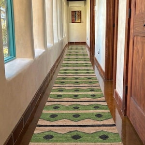 Può includere: Un lungo tappeto rettangolare verde e beige con un motivo geometrico, posto in un corridoio. Il tappeto presenta strisce alternate di verde, beige e forme di diamanti neri. Il testo "Govind Art Rugs" è visibile in basso.