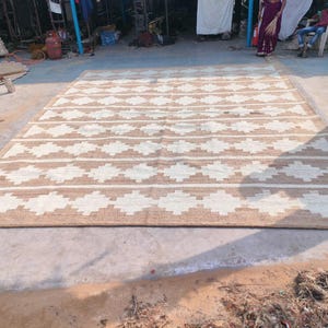 May include: A large, rectangular, woven rug with a geometric pattern. The rug is made of natural fibres and has a light brown and white colour scheme.