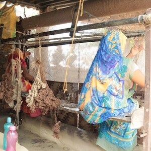 May include: Two people are weaving on a loom in a rustic setting. The loom is made of wood and metal and has a white fabric stretched across it. The people are wearing colorful clothing and are using their hands to weave the fabric.