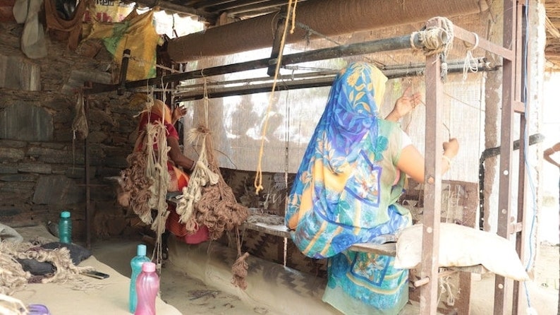 May include: Two people are weaving on a loom in a rustic setting. The loom is made of wood and metal and has a white fabric stretched across it. The people are wearing colorful clothing and are using their hands to weave the fabric.