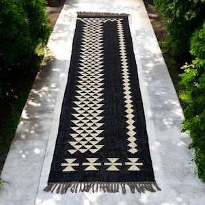 May include: Black and white geometric patterned runner rug with fringe on the ends. The rug is laid out on a light gray concrete surface.