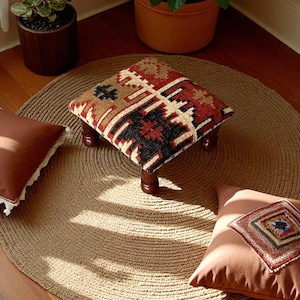 Pode incluir: Um pufe quadrado com um tampo estampado em vermelho, preto e bege está sobre um tapete redondo de juta tecida. O pufe tem quatro pernas curtas de madeira escura. Duas almofadas marrons estão no chão.