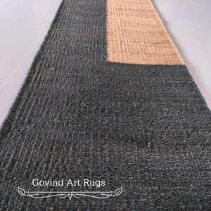 May include: A long, rectangular rug with a dark blue and natural jute design. The rug has a woven texture and a fringe border. The text "Govind Art Rugs" is printed on the rug.