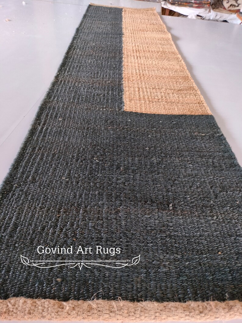 May include: A long, rectangular rug with a dark blue and natural jute design. The rug has a woven texture and a fringe border. The text "Govind Art Rugs" is printed on the rug.