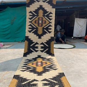 Puede incluir: Una alfombra rectangular larga con un patr&oacute;n geom&eacute;trico en negro, blanco y dorado. La alfombra tiene flecos en la parte superior e inferior.
