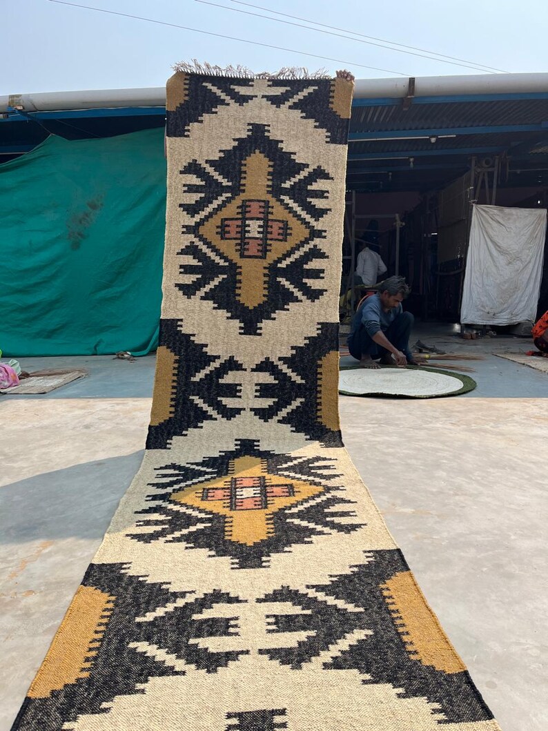 Puede incluir: Una alfombra rectangular larga con un patr&oacute;n geom&eacute;trico en negro, blanco y dorado. La alfombra tiene flecos en la parte superior e inferior.