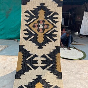 Puede incluir: Una alfombra tejida a mano con un patr&oacute;n geom&eacute;trico en negro, blanco, amarillo y naranja. La alfombra tiene un borde con flecos y est&aacute; hecha de fibras naturales.