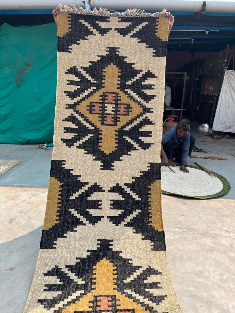 Puede incluir: Una alfombra tejida a mano con un patr&oacute;n geom&eacute;trico en negro, blanco, amarillo y naranja. La alfombra tiene un borde con flecos y est&aacute; hecha de fibras naturales.