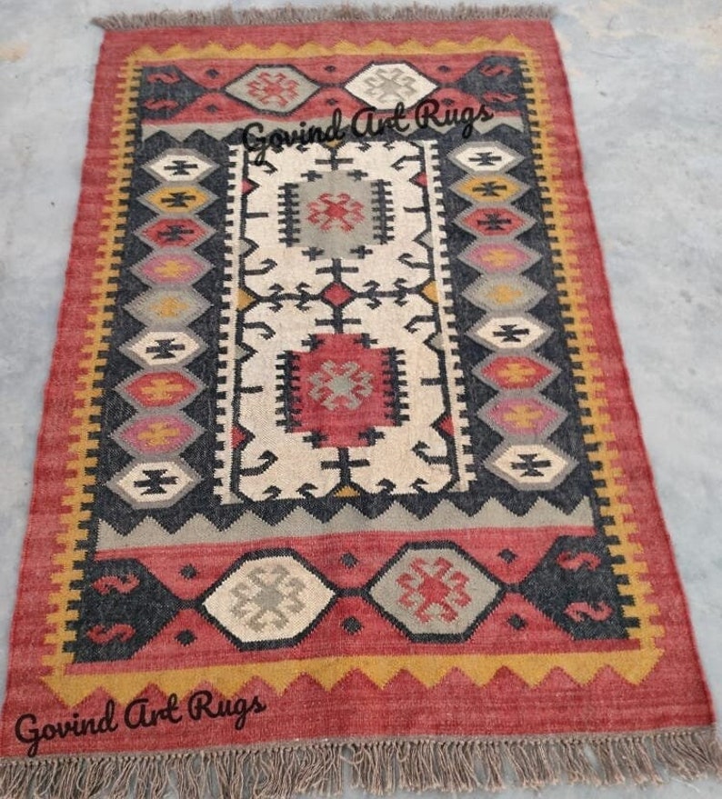 May include: A rectangular rug with a geometric pattern in shades of red, black, white, yellow, and gray. The rug has a fringe border and the text "Govind Art Rugs" is printed on the rug.
