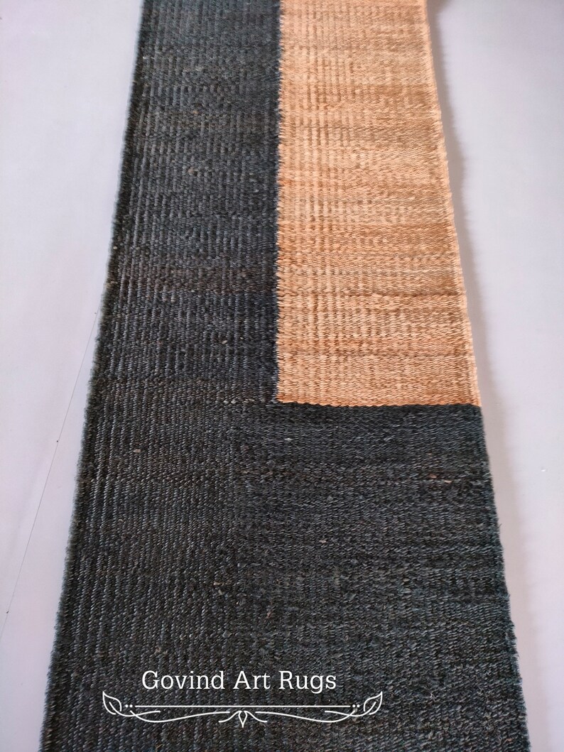 May include: A long, narrow rug with a dark blue and beige color scheme. The rug has a simple geometric design with a rectangular shape in the center. The rug is made of natural fibers and has a textured surface. The text "Govind Art Rugs" is visible at the bottom of the rug.